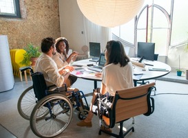 Drie jongeren waarvan een in een rolstoel om een ronde tafel waarop laptop en papieren